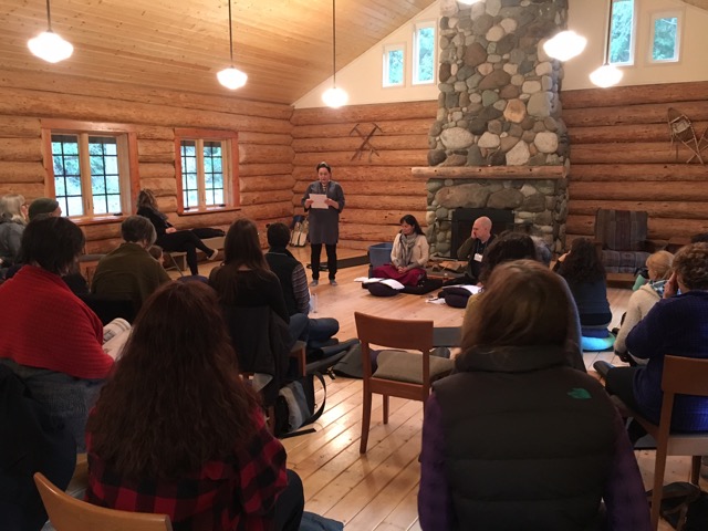 Several mindfulness teachers sit in front of a large stone fireplace in a tall bright log room, with about a dozen students facing them.