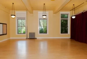 The room that our classes take place in at Good Shepherd in Seattle. High cielings, pale wood floor, three hanging overhead lights, yellow walls, and three tall windows with white frames and greenery outside.