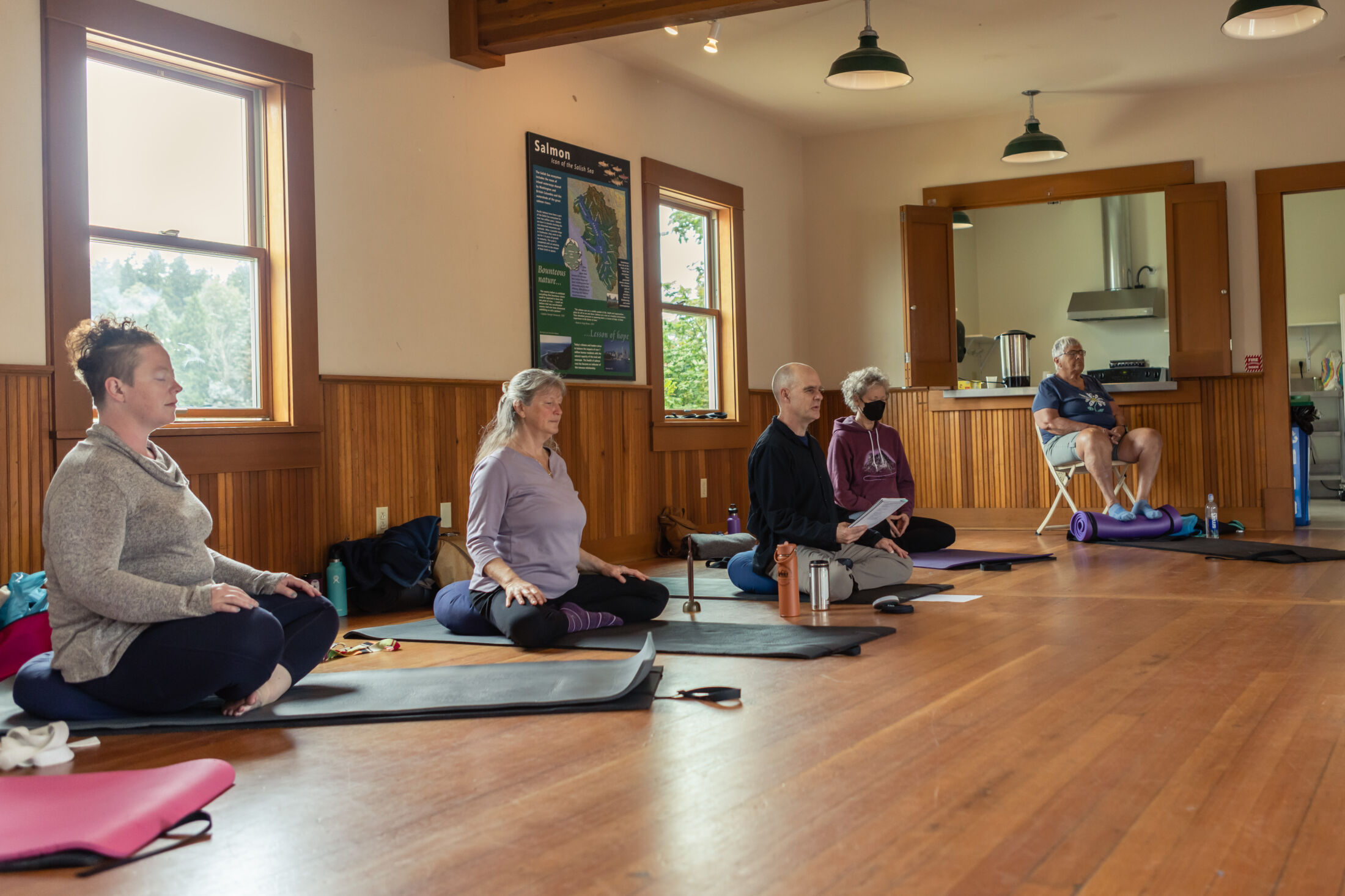 Meditators in a brightly lit room on a hardwood floor, sitting on cushions and yoga mats meditating. 