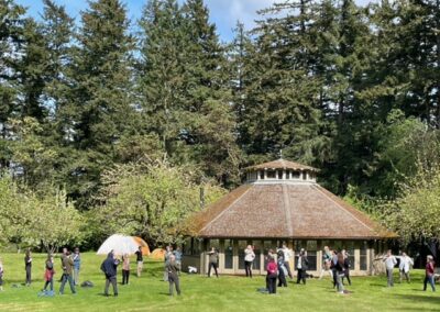 A photo of a large pavilion in an apple orchard, with dozens of people standing outside it practicing mindful movement in the sunshine. Pine trees in the background and a hint of blue sky.