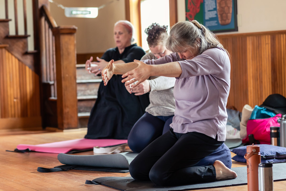 Mindful movement instructor Raizelah Bayen and several participants next to her kneel on a yoga mat and bow their heads in a light wooden room. Raizelah is in a purple shirt and black yoga pants.