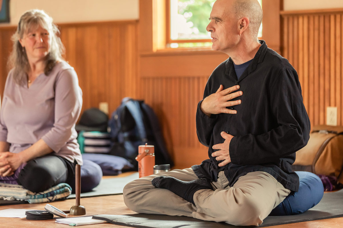 Tim Burnett, Lead instructor, sits with legs crossed and his hands on his torso as he explains something with deep feeling. Tim's wearing a black button down shirt and khakis,