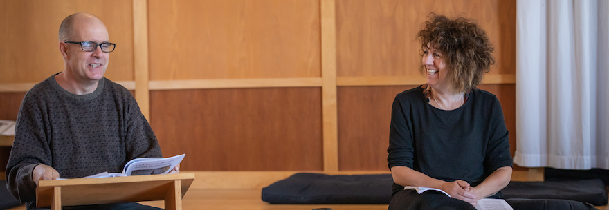 Head instructor Tim Burnett sits on the floor of a bright wooden room, reading from his text and laughing with class participants.