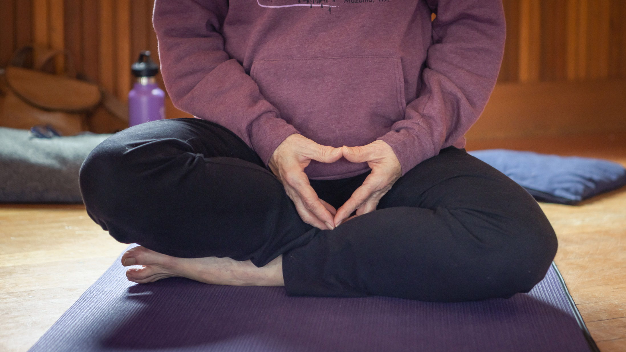 The crossed legs and balanced hands of a person sitting on a purple yoga mat.