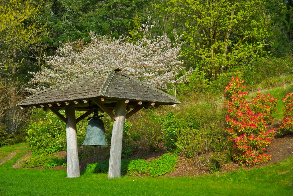 A large blue-green bell hangs in an awning amongst the flowering garden bushes at Whidbey Institute.