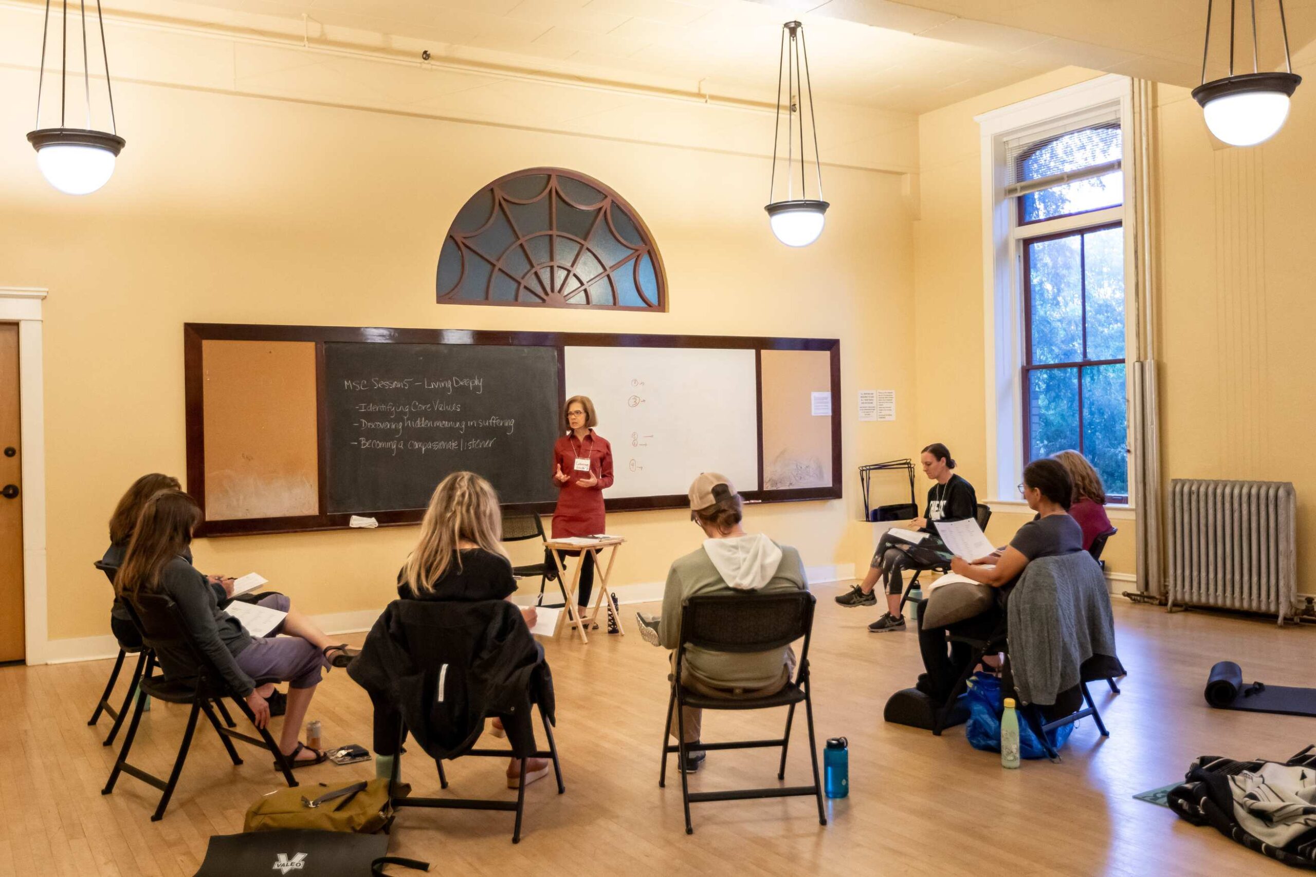 A bright room with high ceilings that has a circle of chairs facing a chalkboard, where the teacher stands and teaches mindfulness topics.