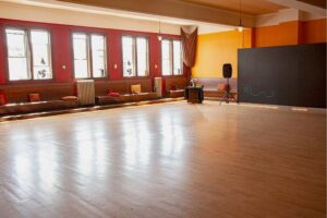 A large wooden dancefloor in a big room with bright natural light coming through many tall windows.