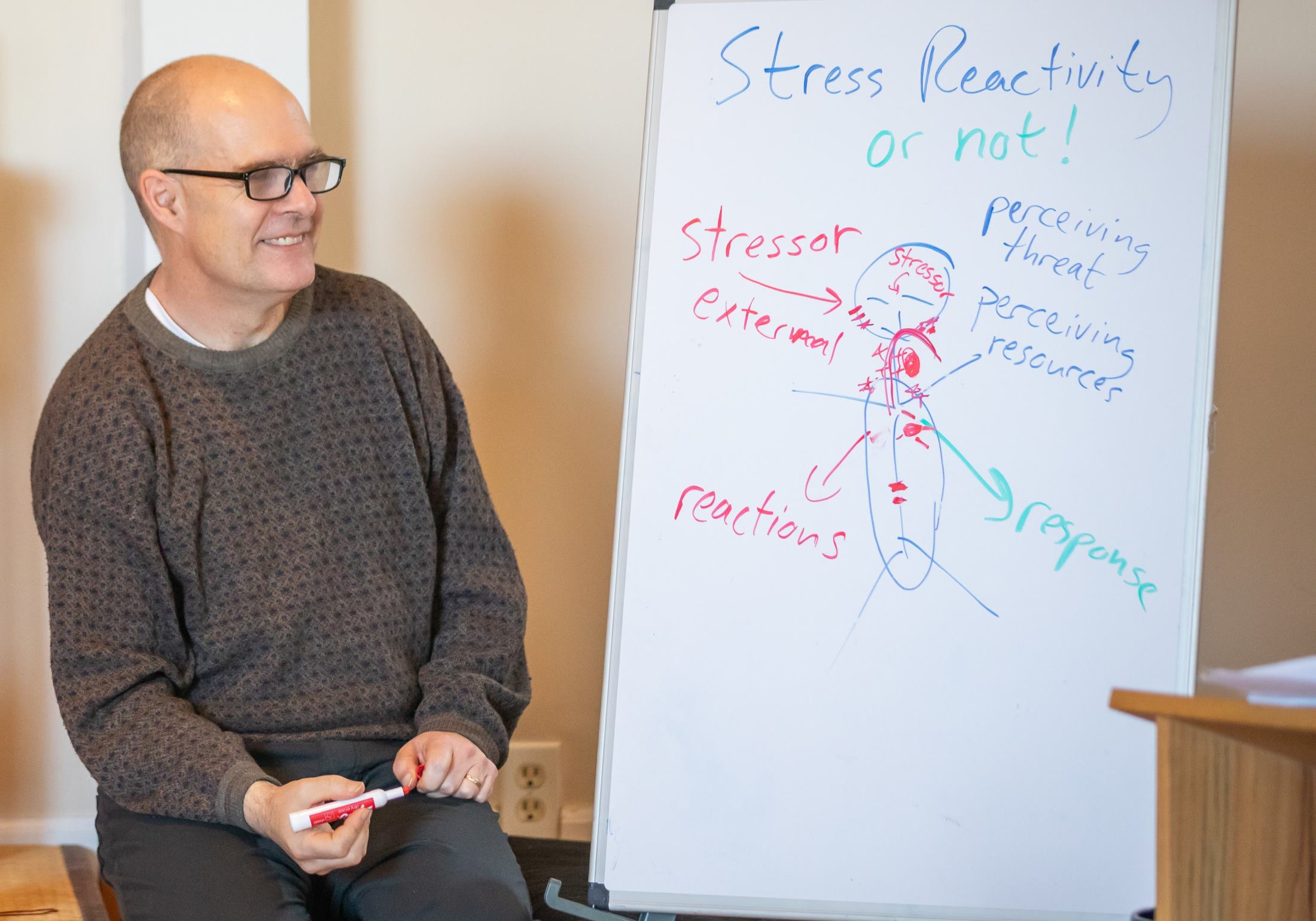 Teacher Tim Burnett kneels next to a whiteboard, with a diagram of a human body and text describing the stress response.